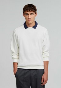 Polo alb cu mâneci lungi și guler bleumarin, având o croială relaxată și o textură moale. Se combină cu pantaloni gri închis.