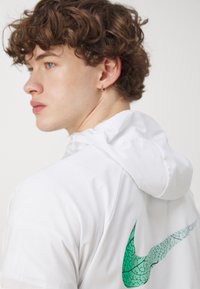 Biela kapucňa má na chrbte textúrovaný zelený znak Nike. Tkanina vyzerá ľahká a má hladkú textúru.