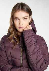 Lilla puffjakke med hætte, der har lynlåslukning og synligt quiltet mønster. Stoffet ser glat ud og er isoleret.
