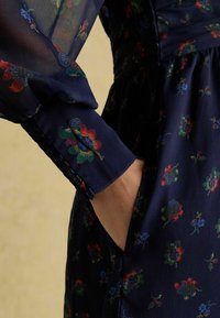 Merine lilleline kleit, millel on läbipaistev kangas ja värvilised lillemustrid. Varrukal on kogumisse detail, ja käsi on asetatud küljetaskusse.