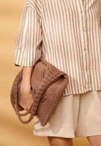 Personne portant un short beige et une chemise à rayures boutonnée tenant un sac à main marron tricoté avec des poignées tressées sur un fond beige.