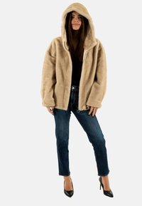 Giacca cappotto in finta pelliccia beige con vestibilità ampia, chiusura con zip e tasche laterali, abbinata a jeans blu scuro e tacchi alti neri.