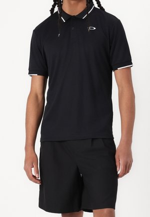 Homme portant un polo noir à bordures blanches et un short noir, debout devant un fond uni.