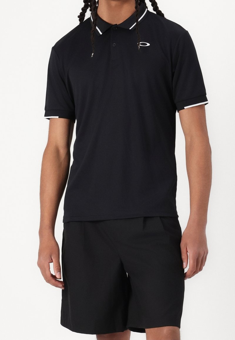 Homme portant un polo noir à bordures blanches et un short noir, debout devant un fond uni.