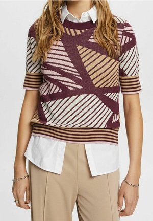 Vrouw draagt een trui met korte mouwen met geometrische strepen in bordeaux, beige en wit over een wit overhemd met kraag en een beige broek.