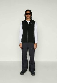 Colete preto com fecho de correr sobre uma camisa de mangas longas branca, combinado com calças pretas com bolsos com fecho e calçado negro.