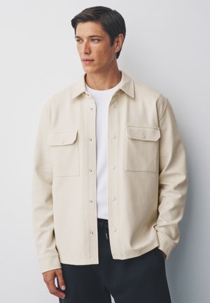 Tânăr cu păr scurt și întunecat, purtând o jachetă bej cu nasturi peste o cămașă albă, stând în fața unui perete simplu și deschis, privind spre stânga.