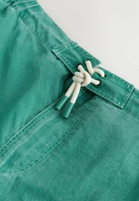 Short en tissu vert avec cordon blanc noué en nœud à travers des œillets verts à la taille sur fond blanc.