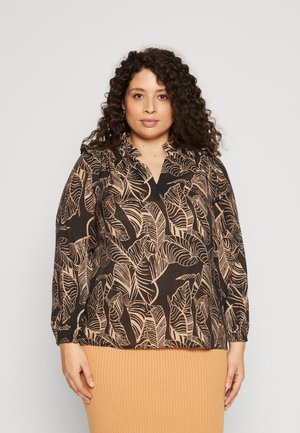 Blouse noire avec un motif de feuilles beige clair, décolleté en V et manches longues. Tissu texturé avec des poignets froncés, portée sur une jupe beige à côtes.