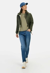 Groene pufferjack met horizontale stiksels, olijfgroene t-shirt, blauwe spijkerbroek en witte sneakers, geaccessoreerd met een blauwe pet.