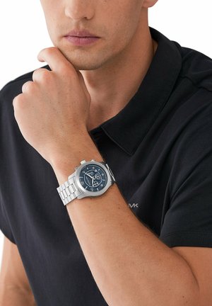 Reloj de acero inoxidable plateado con una esfera azul redonda, números blancos y una pulsera de eslabones, llevado en una muñeca con una mano descansando sobre la barbilla.