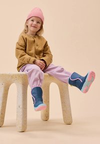 Blaue Gummistiefel mit rosa Akzenten, lilafarbene Hose und eine hellbraune Hemdjacke. Ein Kind sitzt auf einem strukturierten, hellen Hocker.