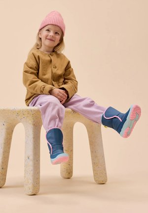 Blue rain boots with pink accents, lilac pants, and a tan button-down jacket. Child sits on a textured, light-colored stool.