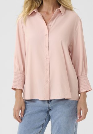 Vrouw draagt een lichtroze blouse met knopen en geplooide manchetten en lichtblauwe spijkerbroek, staand tegen een effen achtergrond.