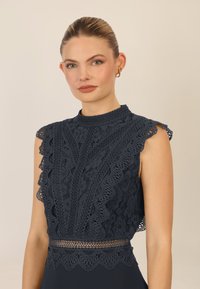Top in pizzo blu navy con bordi ondulati, collo alto e motivo floreale intricati. I dettagli trasparenti in vita aggiungono texture e contrasto.