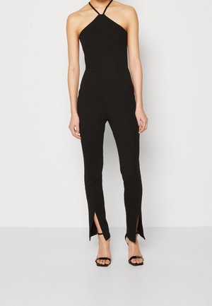 Kobieta ubrana w czarny kombinezon na szyję z rozcięciami przy kostkach, w połączeniu z czarnymi sandałami na wysokim obcasie na cienkich paskach, na jednolitym tle.