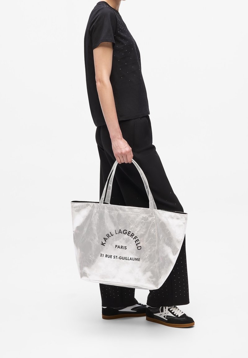 Personne en tenue noire tenant un grand sac cabas argenté Karl Lagerfeld avec texte noir, debout sur fond blanc uni.