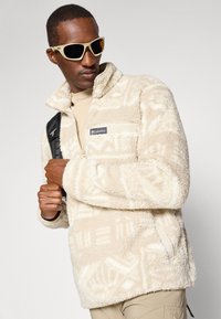 Beige fleecový pulovr s geometrickým vzorem, zapínáním na zip v předním kapse a vysokým límcem. Obsahuje těsně přiléhající manžety a boční kapsy.