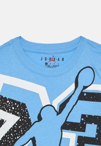 Ανοιχτό μπλε βαμβακερό μπλουζάκι με μαύρη φιγούρα καλαθοσφαιριστή, τοποθετημένη σε ένα τολμηρό γεωμετρικό σχέδιο με λευκές και μαύρες λεπτομέρειες.