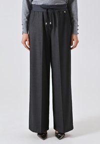 Donna che indossa pantaloni larghi grigi scuri con vita regolabile e un top grigio a maniche lunghe, in piedi con le mani rilassate lungo i fianchi.