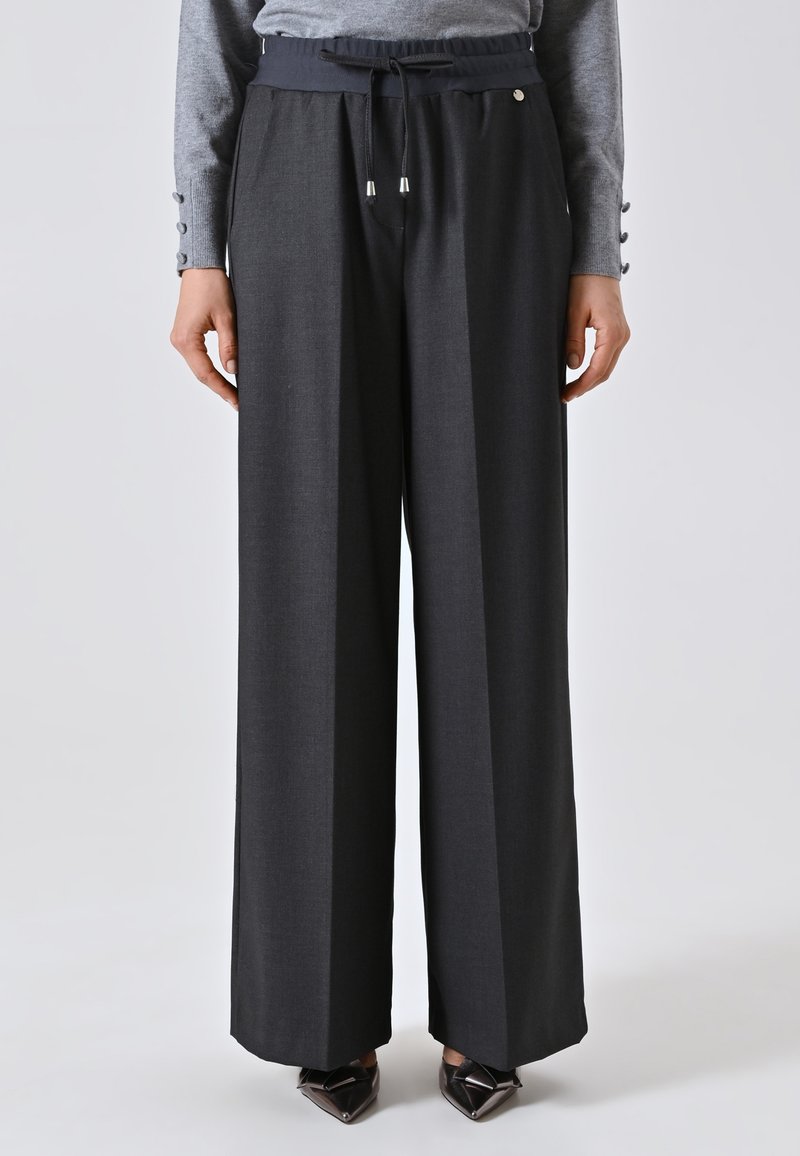 Donna che indossa pantaloni larghi grigi scuri con vita regolabile e un top grigio a maniche lunghe, in piedi con le mani rilassate lungo i fianchi.