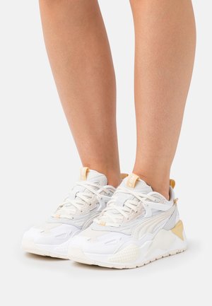 White and beige athletic sneakers worn on bare legs, shown from the front and slightly to the side, on a white background.
