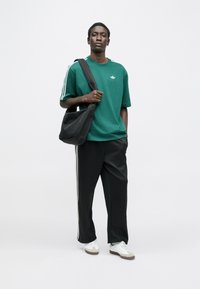 Zelené bavlnené tričko s bielym logom, spojené s čiernymi nohavicami s bielymi bočnými pruhmi. Modelka nesie čiernu tašku cez rameno.