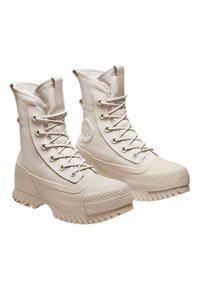 Botas blancas con cordones, suela de goma, detalles texturizados en la punta y parte superior de tela. Presenta un logo redondo en el lateral.