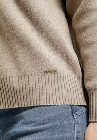 Beżowy sweter z dzianiny z prążkowanym wykończeniem u dołu i przy mankietach. Posiada małą metalową metkę z logo na dole. Nosi go się na niebieskim dżinsowym materiale.