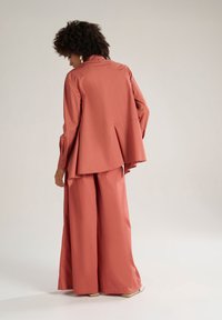 Giacca oversize color corallo e pantaloni a gamba larga in stoffa leggera, con una piega sul retro e polsini abbottonati per un tocco di dettaglio.