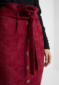 Röd mockaskirt med knyt i midjan, med guldknappar fram. Nära vy av textur och detaljer, med en svart topp synlig.