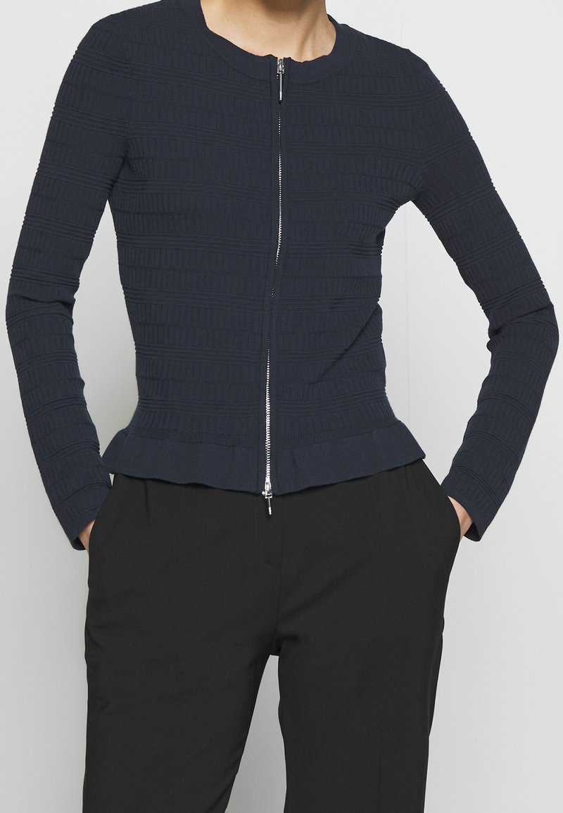 Femme portant une veste zippée texturée bleu marine ajustée et un pantalon noir, les mains dans les poches, debout devant un fond clair uni.