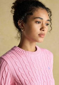 Młoda kobieta z kręconymi włosami w różowym swetrze w warkocze, nosząca złote kwadratowe kolczyki, patrząca z zamyśleniem w bok na beżowym tle.