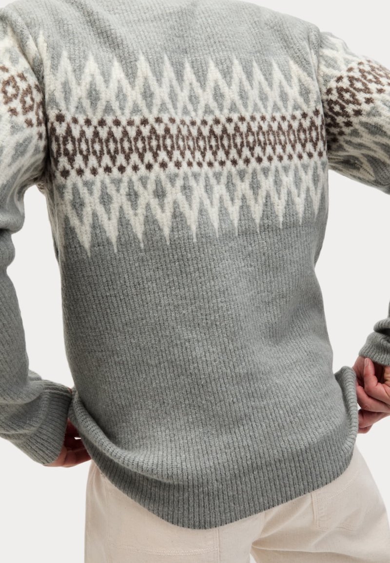 Grijze gebreide trui met witte en bruine geometrische patronen over de borst en mouwen, gedragen door een persoon die de zoom aanpast.
