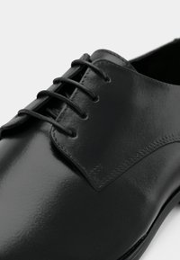 Schwarzer Lederschuh mit polierter Oberfläche, ausgestattet mit sechs Ösen, flachen Schnürsenkeln und einem genähten Kapuze-Design. Glatte Textur und schlanke Silhouette.