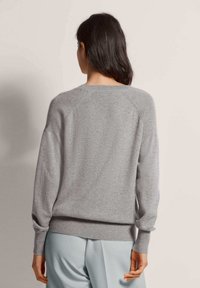 Frau mit langen dunklen Haaren, die einen hellgrauen gestrickten Pullover und eine hellblaue Hose trägt, mit dem Rücken zu einer schlichten Hintergrund. 