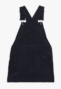 Tmavě modré manšestrové šaty na traky s nastavitelnými popruhy, dvěma předními kapsami a texturovaným žebrovaným vzorem. Doplněné kováním ve stříbrném tónu.