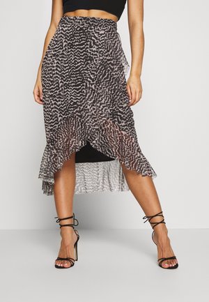 Femme portant une jupe midi à motif abstrait noir et blanc, ourlet asymétrique à volants, associée à des sandales noires à talons hauts et à lanières.