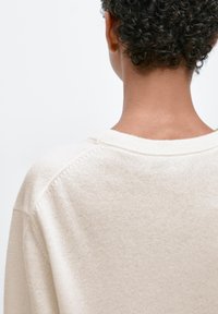 Ljusbeige tröja med vid halsringning och ribbade kanter, med en mjuk, texturerad stickning. Baksidan visar en avslappnad passform och sömdetaljer.