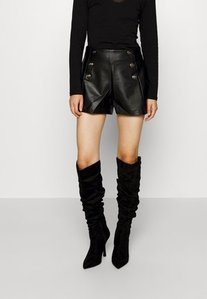 Black leather high-waisted shorts with front pleats and decorative silver buttons, paired with knee-high, slouchy black suede boots.