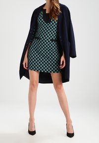 Zwarte jas over een meerkleuren jurk met een geometrisch patroon in teal en wit. Hoge hakken maken de outfit compleet.