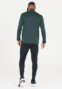 Langemouw, donkergroen sporttop met een ritssluiting aan de kraag en een gestructureerd patroon, gecombineerd met zwarte, strakke leggings en sportschoenen.