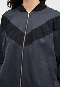 Temno siva bomber jakna z črnim nabranim rjuhastim delom na ramenih, srebrnim zadrgo, rebrastim ovratnikom in stranskimi žepi. Gladek material z mat zaključkom.