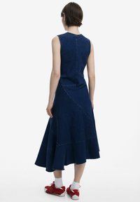 Jeansklänning i mörkblått med ärmlös design. Har kontrasterande sömdetaljer, utsvängd kjol och längd till mitten av vaden. Kombineras med röda sneakers.