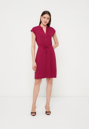 Kvinde stående mod hvid baggrund iført en magentarød knælængdekjole med korte ærmer, knapper og taljebånd samt sorte sko med åben tå.