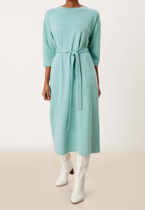 Femme portant une robe midi bleu pastel à manches longues avec une ceinture à la taille et des bottes blanches à talons montant jusqu'aux genoux, debout sur un sol beige.