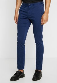 Man med slim-fit marinblå chinos och svarta snörskor, stående mot en enkel ljus bakgrund.