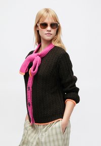 Tmavohnedý pletený sveter s jasne ružovým golierom a gombíkmi, ozdobený oranžovými akcentmi. Model má na sebe slnečné okuliare a nohavice s potlačou.