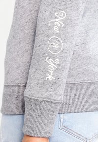 Grijze sweatshirt met een gestructureerde afwerking, voorzien van witte tekst "New York" op de mouw en geribde boorden. Het ontwerp bevat een circulair logo.