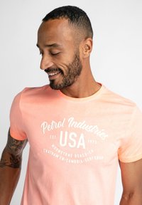 Srajca s kratkimi rokavi v svetlo roza barvi, izdelana iz mehkega blaga, z belim vintage logotipom in besedilom, krojnim ovratnikom in rahlo teksturo.